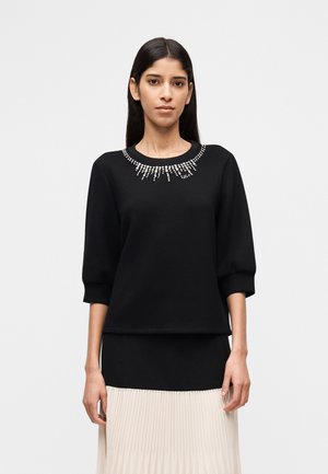 Kvinde iført en sort langærmet top med rhinstenudsmykninger omkring halsudskæringen og et plisseret beige skørt.