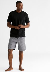 Zwarte katoenen t-shirt met korte mouwen, gecombineerd met knielange zwart-wit geblokte shorts, staand op blote voeten tegen een neutrale achtergrond.