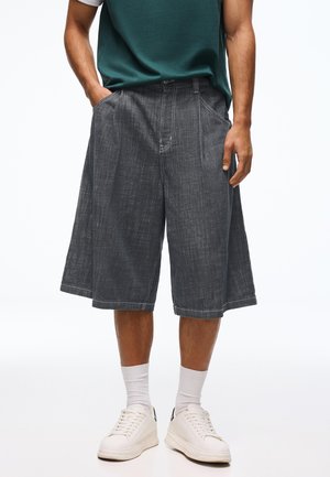 Man wearing loose, knee-length dark gray denim shorts, white socks, white sneakers, and a dark green short-sleeve shirt with hands in pockets.