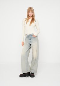 Šviesiai beige, rauktas ilgomis rankovėmis marškinėliai, derinami su išblukusiais mėlynais plačiais džinsais. Juodi masyvūs batai su storu padu užbaigia įvaizdį.