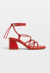 Sandale à talon rouge avec un talon bloc épais, des lanières fines et des liens à la cheville. Texture lisse avec des accents noués. Design à bout ouvert.