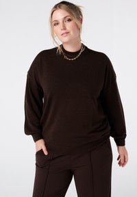 Bruine gebreide trui met een ontspannen pasvorm, verlaagde schouders en geribbelde manchetten en zoom. Gecombineerd met donkerbruine broek. Gouden ketting.