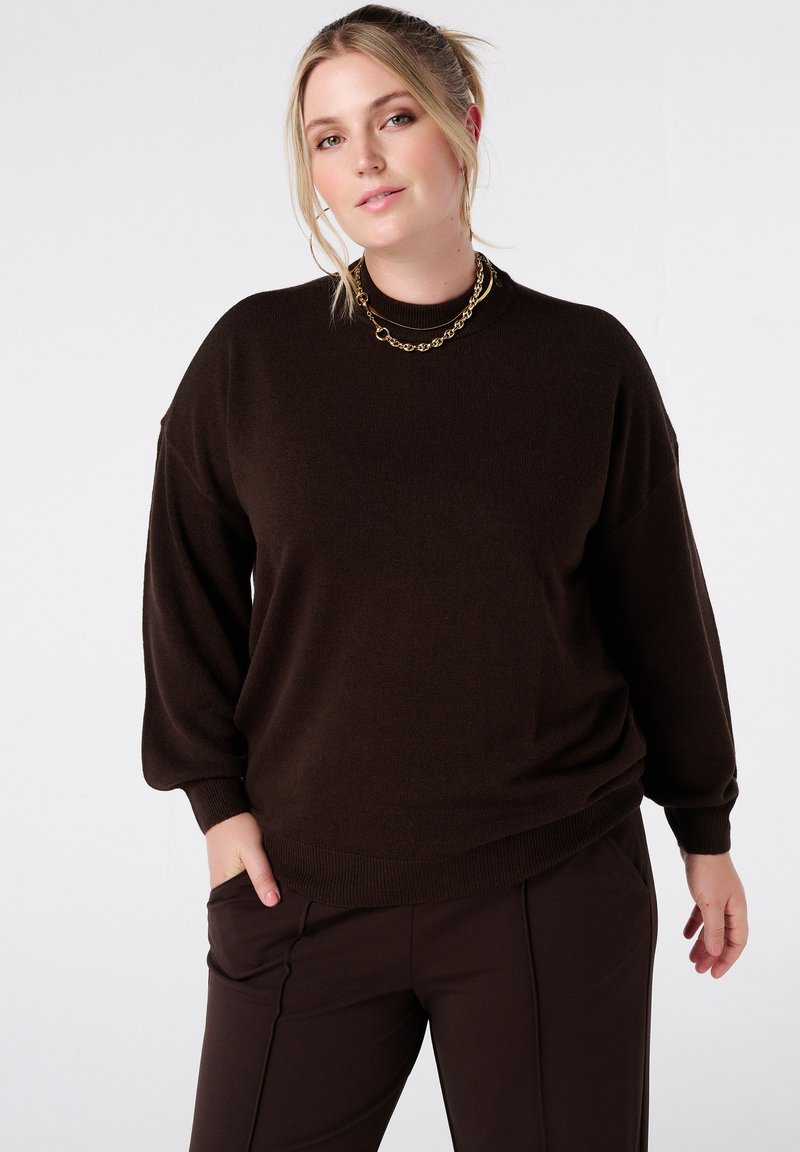 Bruine gebreide trui met een ontspannen pasvorm, verlaagde schouders en geribbelde manchetten en zoom. Gecombineerd met donkerbruine broek. Gouden ketting.