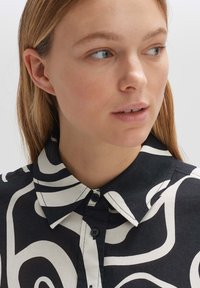 Camicia bianca e nera con motivi astratti, dotata di colletto, chiusura con bottoni e vestibilità comoda. Texture del tessuto liscia e visibile.