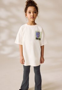 T-shirt en coton blanc avec un motif floral vert et violet et un texte noir sur la poitrine, associé à un pantalon gris ajusté.