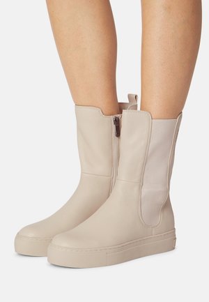 Stivaletti alla caviglia in pelle beige con una texture liscia, cerniere laterali e una spessa suola platform bianca. Presentano un design con ritaglio nella parte superiore.