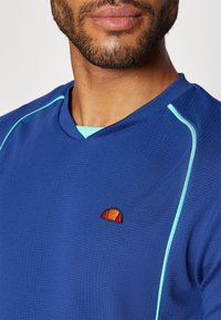 Cămașă sportivă pentru bărbați, de culoare albastră, cu un material texturat. Are accente turcoaz și un mic logo colorat pe piept. Design cu decolteu în V.