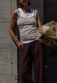 Person wearing sunglasses, beige tank top over white shirt and dark pants, holding a bouquet of small white flowers wrapped in brown paper.