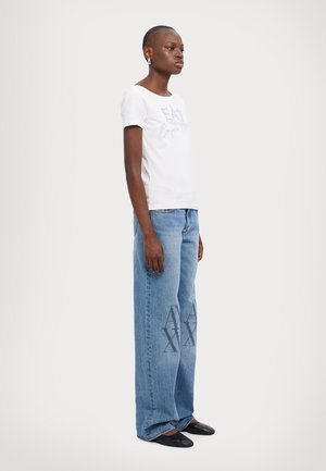 Camiseta blanca de manga corta con estampado gráfico gris, combinada con jeans de pierna ancha azul claro que tienen un patrón decorativo azul oscuro en las piernas.