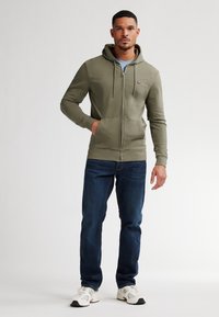 Felpa con zip verde oliva, dotata di tasca frontale e cappuccio con coulisse, abbinata a jeans blu scuro e sneakers bianche. Texture liscia.