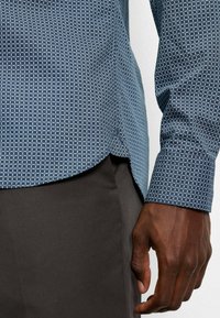 Blaues gemustertes Hemd mit geometrischem Design, langen Ärmeln, Knopfmanschetten und abgerundetem Saum, kombiniert mit dunklen Hosen.