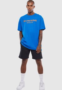 Blå bomulds T-shirt med orange tekst, der står "livet er en daglig kamp", parret med sorte shorts og hvide sneakers. Korte ærmer, afslappet pasform.