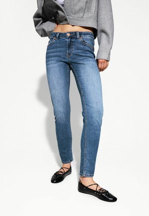 Vrouw draagt hoge taille smalle blauwe jeans, zwarte veterloze platte schoenen en een kort grijs jasje, staand tegen een witte achtergrond.
