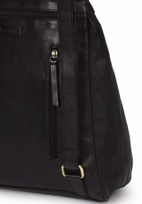 Esquina de una bolsa de cuero negra con bolsillo vertical con cremallera, correa de cuero y hebilla de latón sobre fondo blanco.