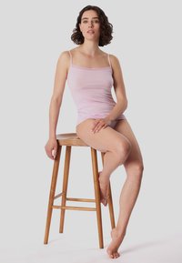 Light pink camisole with thin straps, soft fabric, fitted design. Model sitting on a wooden stool, showcasing the tank top.