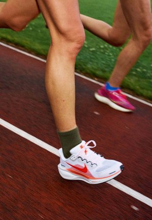 Close-up van de benen en voeten van twee hardlopers in sportschoenen die rennen op een rode atletiekbaan naast groen gras.