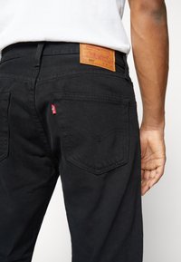 Jeans de denim negro con un parche de cuero en la cinturilla trasera, que cuentan con bolsillos traseros distintos y un detalle de etiqueta roja.