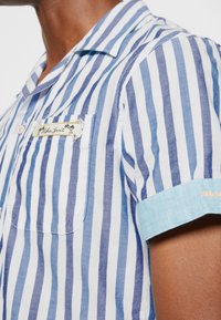 Camisa de manga corta con rayas verticales azules y blancas, un bolsillo parche y un puño azul claro con texto naranja. Confeccionada en tela texturizada.