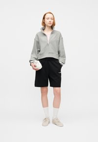 Άτομο με γκρι φούτερ με φερμουάρ στο τεταρτημόριο, μαύρα σορτς Calvin Klein, λευκές κάλτσες, μπεζ αθλητικά παπούτσια, κρατώντας μικρό λευκό τσαντάκι μπροστά σε μονόχρωμο φόντο.