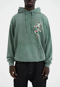 Groene hoodie van zacht fabric met een voork pocket, voorzien van een kleurrijke schedel- en rozenafbeelding. Losse pasvorm met trekkoorden en opgebouwde mouwen.