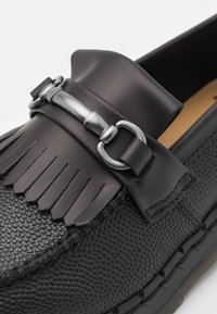 Mocassin en cuir noir à enfiler avec une surface texturée, des détails en frange et un accent de boucle métallique sur le cou-de-pied.