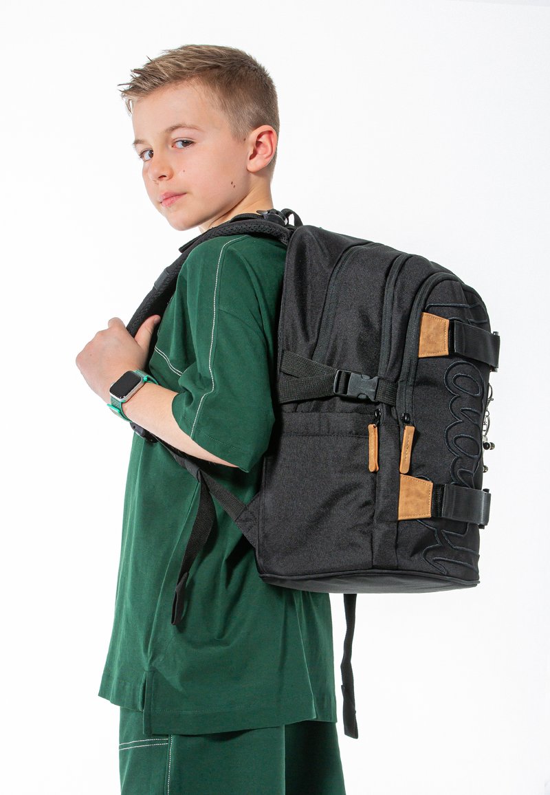 Zaino nero con accenti marroni e logo impresso, dotato di spallacci imbottiti e più scomparti. Il modello indossa una maglietta verde.