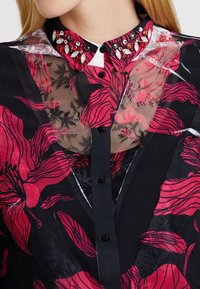 Durchsichtige schwarze Bluse mit lebhaftem roten Blumenmuster, ausgestattet mit einem weißen Kragen, der mit schwarzen und roten Verzierungen sowie schwarzen Knöpfen geschmückt ist.