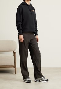 Persoon draagt een zwarte hoodie met klein logo, donkere broek en grijze hardloopschoenen, staand binnen naast een beige fauteuil op een lichte vloer.