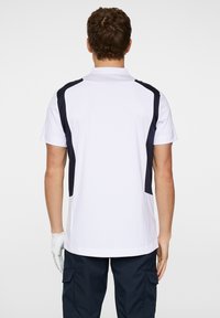 Vit polo skjorta med mörk marinblå sidopaneler, korta ärmar och rak fåll. Bärs över marinblå arbetsbyxor och en vit golfhandske.