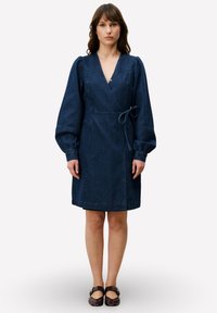 Robe portefeuille en denim avec manches longues bouffantes, décolleté en V profond et ceinture à nouer. Présente des rayures verticales et un ourlet arrivant au genou. Couleur bleu foncé.