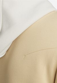 Zbliżenie beżowej tkaniny z białym nakryciem głowy na wierzchu, ukazujące fakturę i szczegóły szycia.