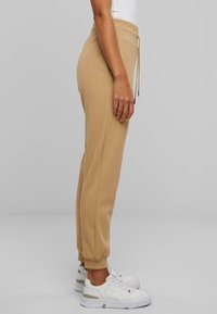 Beige bomullsjoggers med elastiske mansjetter og strikk i midjen, brukt med hvite joggesko. Stoffet ser mykt og glat ut.