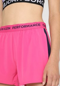 Rosa Sportshorts mit einem schwarz elastischen Bund, auf dem "CALVIN KLEIN PERFORMANCE" steht. Leichter Stoff, gerader Schnitt, seitliche Einsätze in Marineblau.