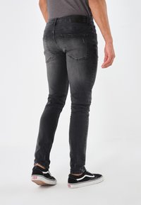 Hombre con jeans negros ajustados y deslavados, y zapatillas Vans negras, de pie contra un fondo blanco.