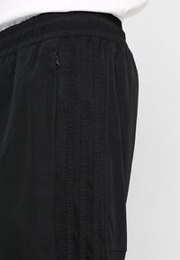 Svart träningsbyxor i lätt tyg med strukturerad randdetalj längs sidorna och dragkedjefickor. Elastisk midja.