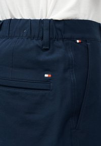 Κοντινό πλάνο σκούρων μπλε παντελονιών με ελαστική μέση και μικρό ορθογώνιο λογότυπο με κόκκινο, άσπρο και μπλε χρώμα κοντά στις πίσω και πλαϊνές τσέπες.