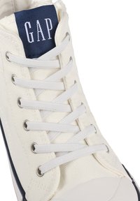 Witte canvas sneakers met witte veters en een marineblauwe textiellabel met "GAP." Rubberen zool met een ronde neus en een minimalistisch ontwerp.