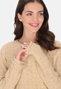 Beige kötött pulóver nyílt mintával, kerek nyakkivágással és hosszú ujjakkal, amelyek hüvelykujjnak nyílásokat biztosítanak, lágy és texturált megjelenést kölcsönözve.