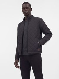 Homme portant une veste matelassée noire par-dessus un pull gris foncé et un pantalon noir, debout avec une main dans la poche sur fond blanc.