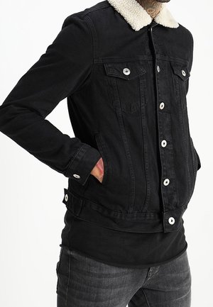 Veste en denim noire avec col en shearling blanc, devant boutonné, poches poitrine et mains dans les poches latérales sur jean foncé.