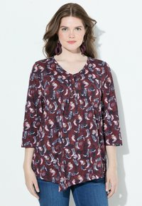 Žena s vlnitými hnědými vlasy, oblečená v vínově zbarvené blouse se vzorem a tříčtvrtečními rukávy, s velkými fialovými náušnicemi, stojící na bílém pozadí.