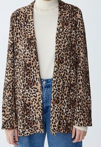 Blazer imprimé léopard en tissu doux, avec devant ouvert, manches longues et coupe ample. Associé à un col roulé de couleur claire.