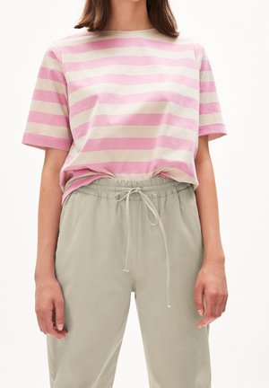 Person trägt ein rosa-beiges T-Shirt mit horizontalen Streifen, in hellgraue Hosen mit Kordelzug gesteckt, steht mit entspannten Armen an den Seiten.