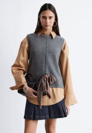 Frau mit langen dunklen Haaren trägt einen grauen, ärmellosen Pullover über einem beigefarbenen Shirt und einem Jeansrock, hält eine schwarze Handtasche mit braunem, gemustertem Schal.