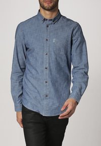 Camisa de mezclilla azul claro con cuello abotonado, botones marrones, mangas largas y un solo bolsillo en el pecho. Textura lisa y corte entallado.