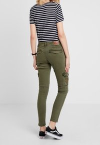 Pantalones cargo ajustados de color verde oliva con bolsillos laterales, detalles en las rodillas y un parche tejido. Combinados con una camiseta de rayas azul marino y blanca.