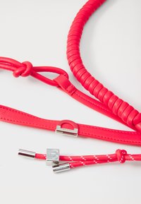 Cinturino in pelle rossa con design intrecciato e sezioni lisce. Presenta dettagli in metallo argentato e ulteriori accenti in corda rossa.