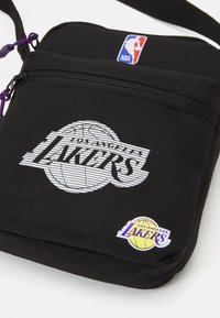 Juodas audinių per petį bagas su baltu tekstu "Los Angeles Lakers". Purpuriniai akcentai ir NBA logotipas viršuje. Uždarymas užtrauktuku su plokščia forma.