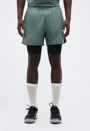 Mees kannab rohelist spordipükste komplekti mustade survelühikute üle, valgeid soonikuga sokke ja musti jooksujalatseid, seistes valge tausta ees.