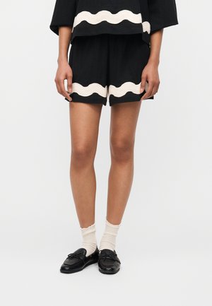 Personne portant un short noir et un haut avec un bord ondulé blanc, des chaussettes crème à volants et des mocassins noirs, debout devant un fond uni.